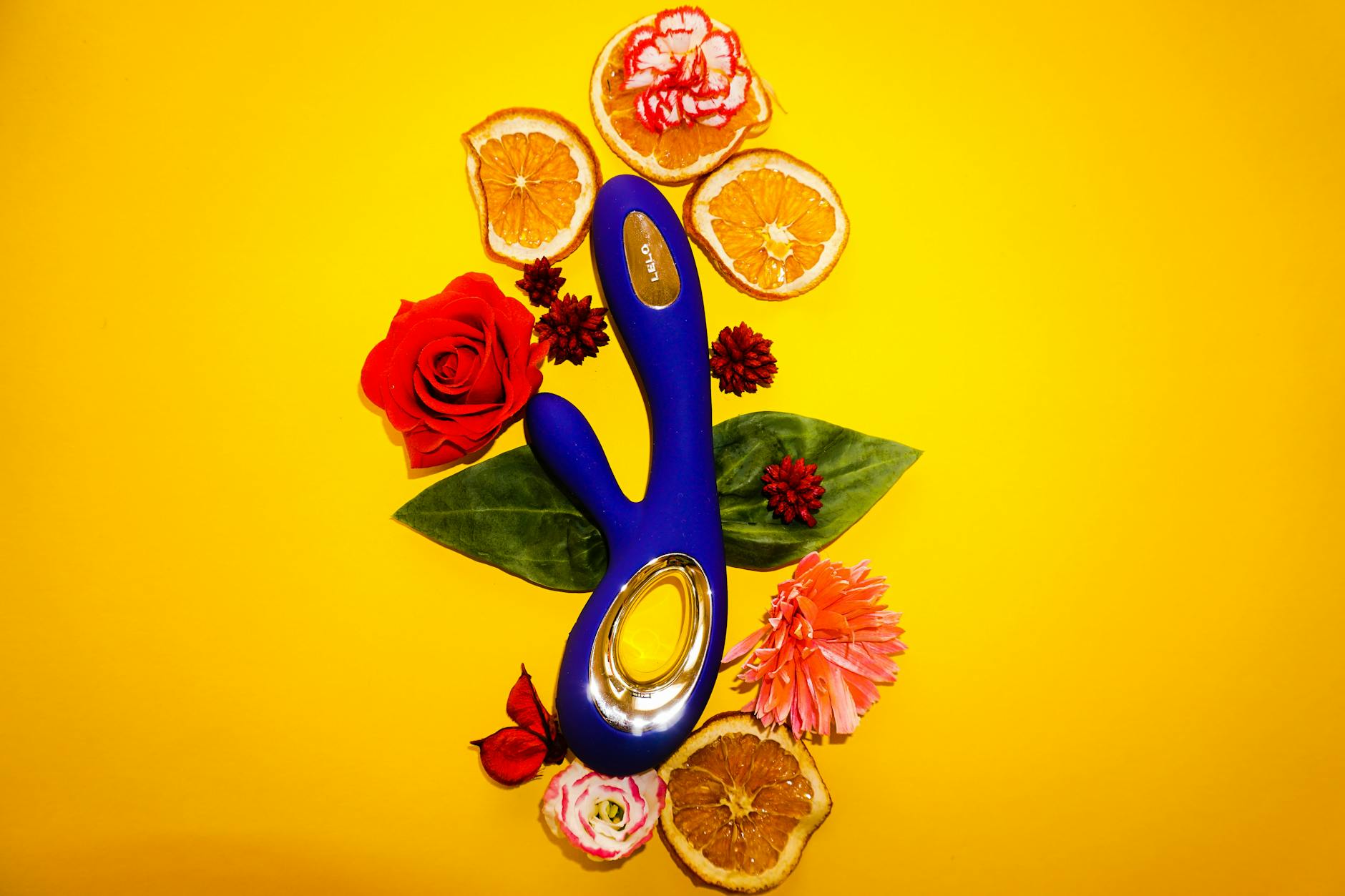 A lemon-yellow vibrator surrounded by flowers and fresh citrus on a bright background, symbolizing sexual wellness throughout your cycle.
